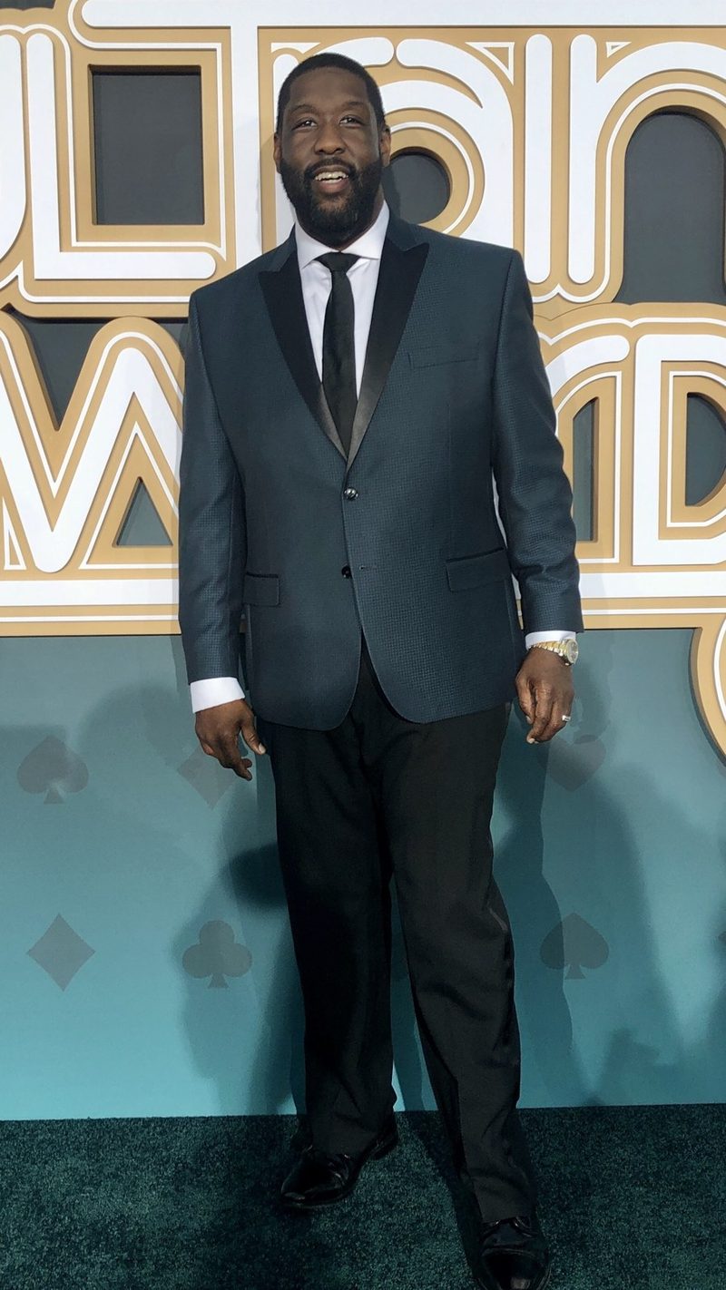 Adam Blackstone in a grey suit and white shirt on a red carpet in front of a gold awards backdrop