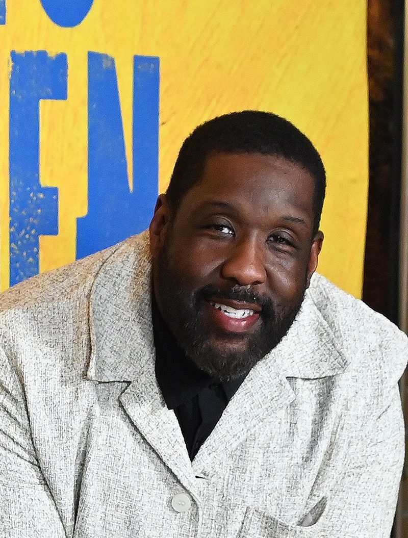 Adam Blackstone smiling in a light grey jacket in front of a bright yellow poster