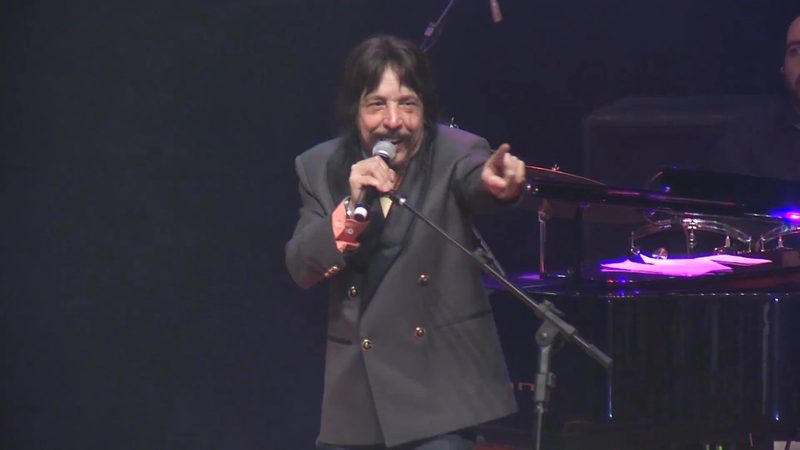 Benito di Paula canta ao microfone em um palco escuro, com um piano ao fundo