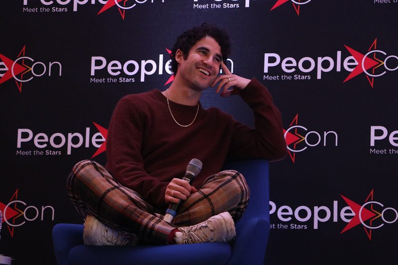 Darren Criss in a burgundy sweater sitting on a stage holding a microphone in front of a PeopleCon backdrop