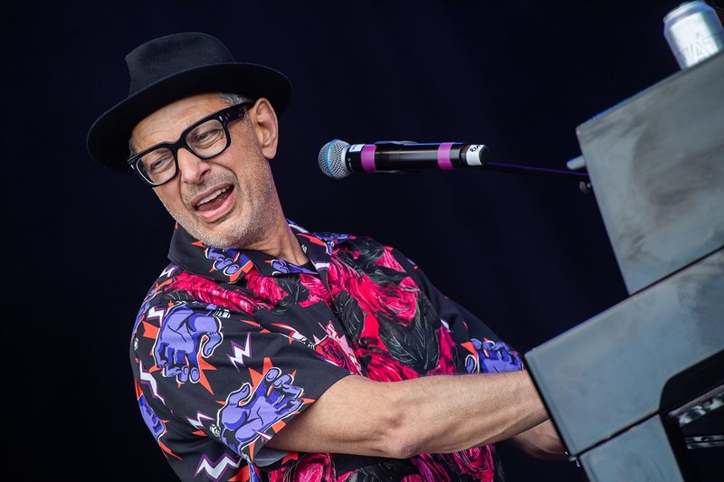 Jeff Goldblum in a hat and patterned shirt playing piano on stage