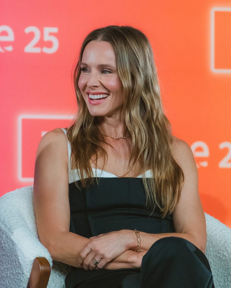 Kristen Bell smiling in a black top in front of an orange backdrop