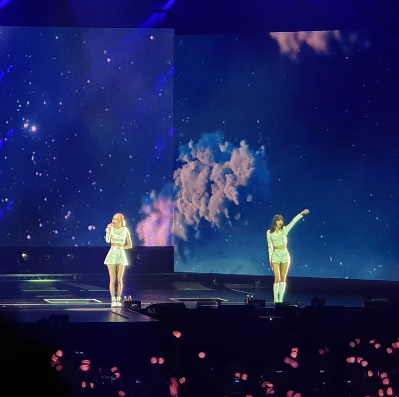 Two performers in matching light outfits on a concert stage in front of a large screen showing a starry sky and pink clouds