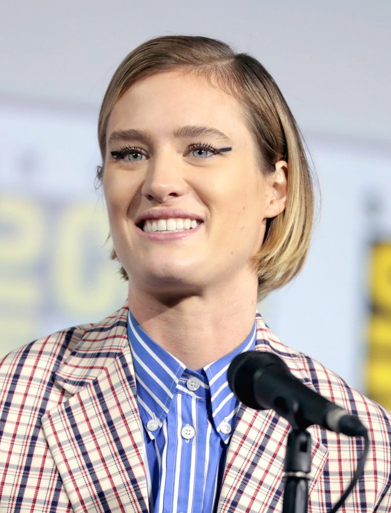 Mackenzie Davis smiling at a microphone in a plaid blazer