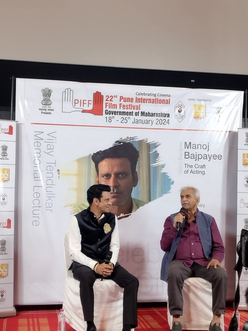 Manoj Bajpayee in a black vest seated on stage beside another man under a Pune International Film Festival banner