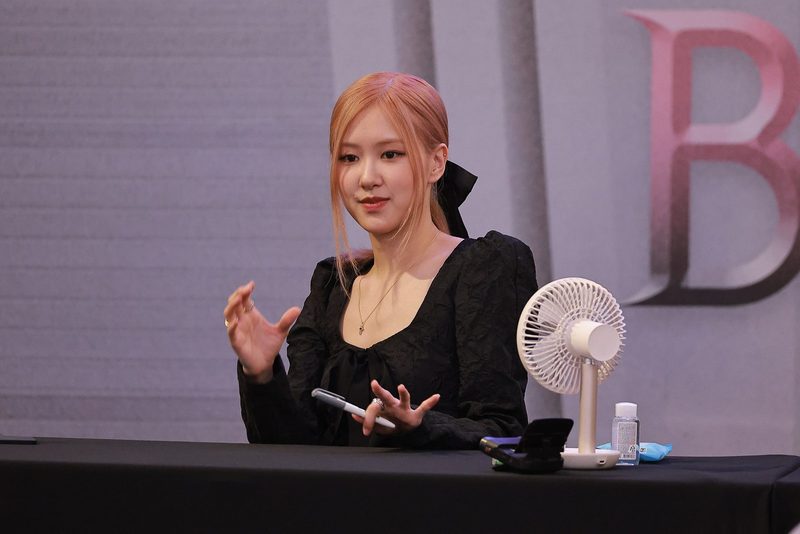 Rosé with pink hair in a black long-sleeved dress, seated at a signing table