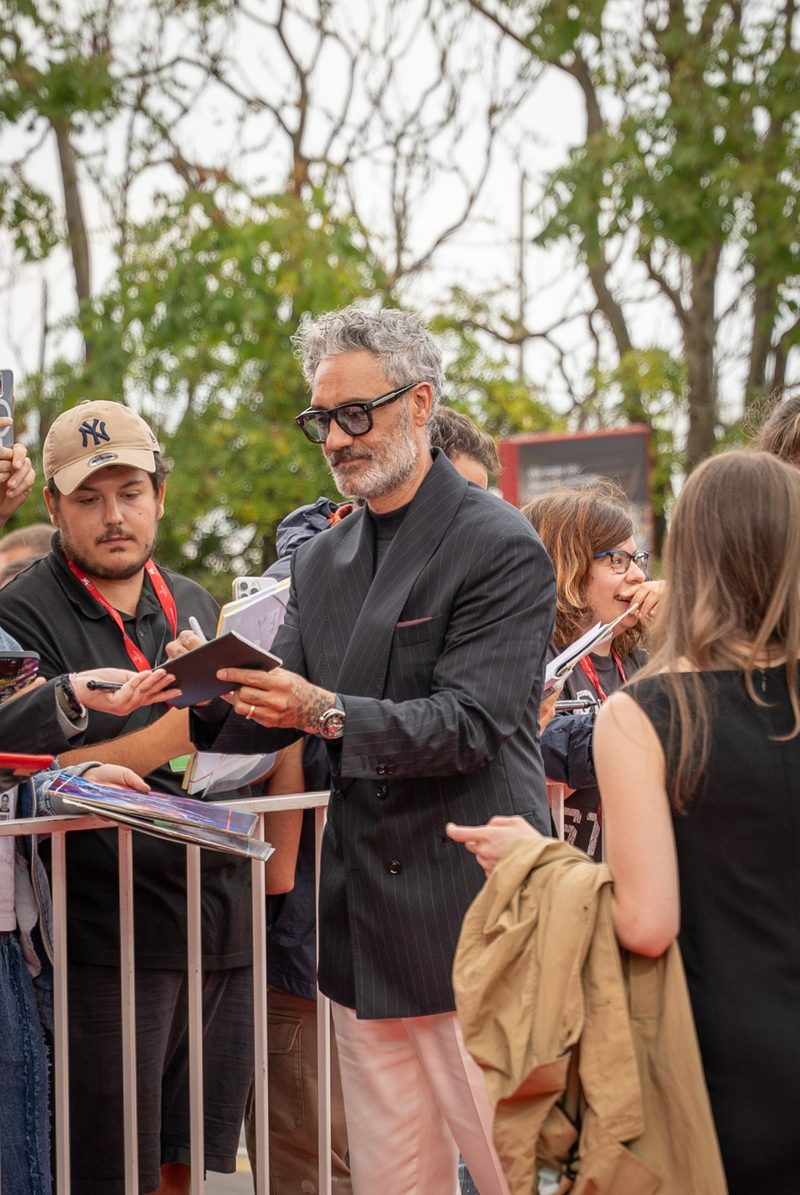 Taika Waititi in glasses and a dark jacket signing autographs for fans