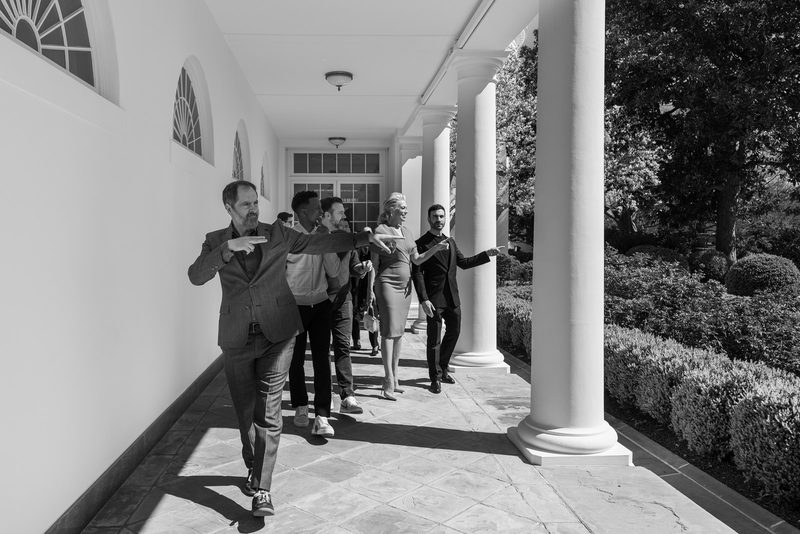 Black-and-white photo of a group of actors walking in a line along a white colonnade beside a garden